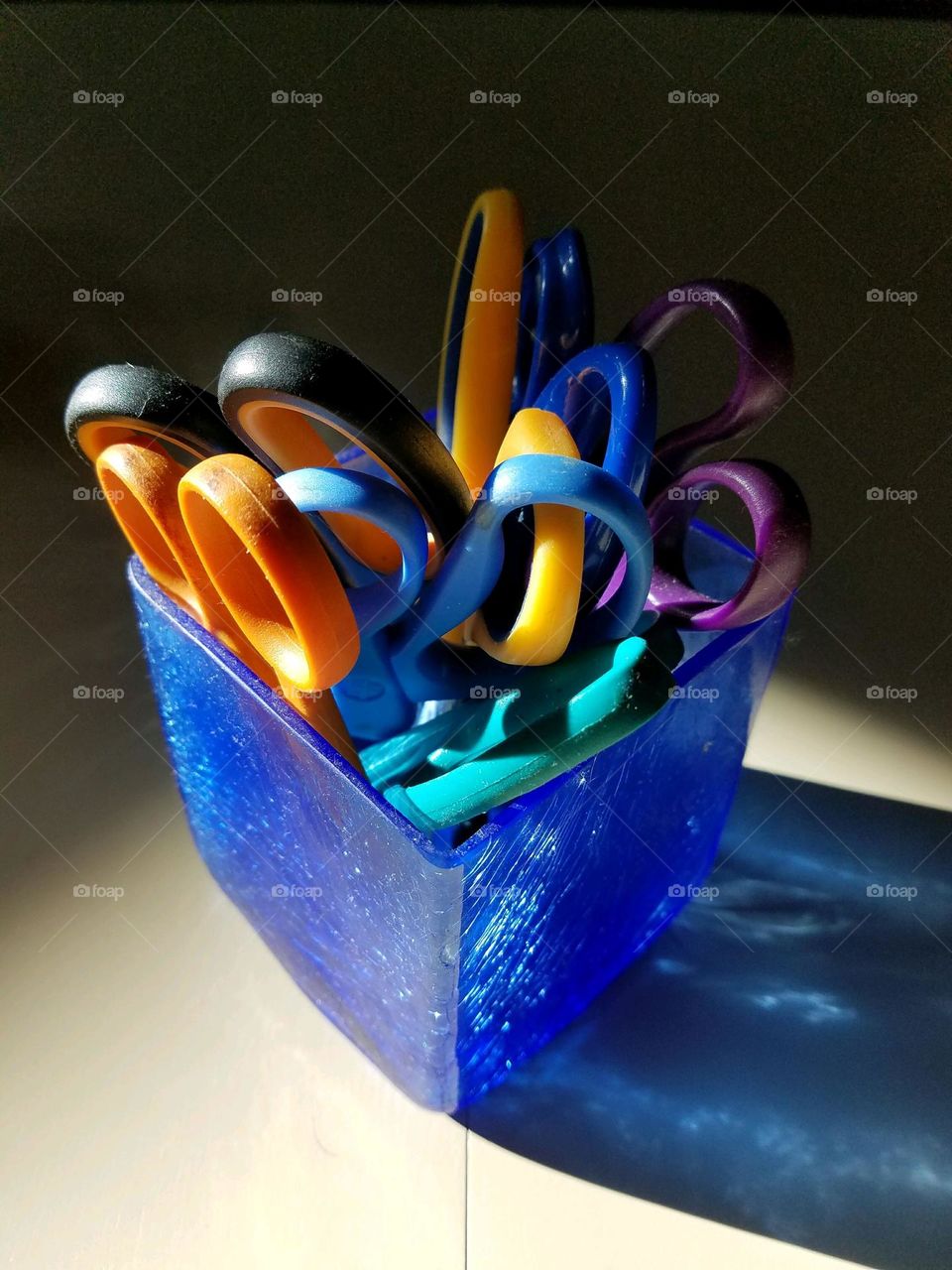Scissors of colors and different sizes clustered in a blue square glass. Scissors are pointed down into holder.