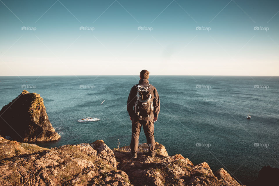 A rear view is male hiker or climber standing on top of a rugged cliff too and looking out to a vast ocean