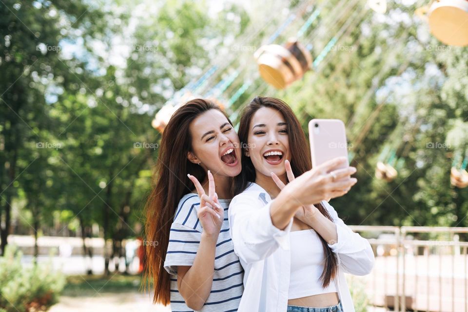 Young women with long hair friends having fun taking selfie on mobile phone at amusement park