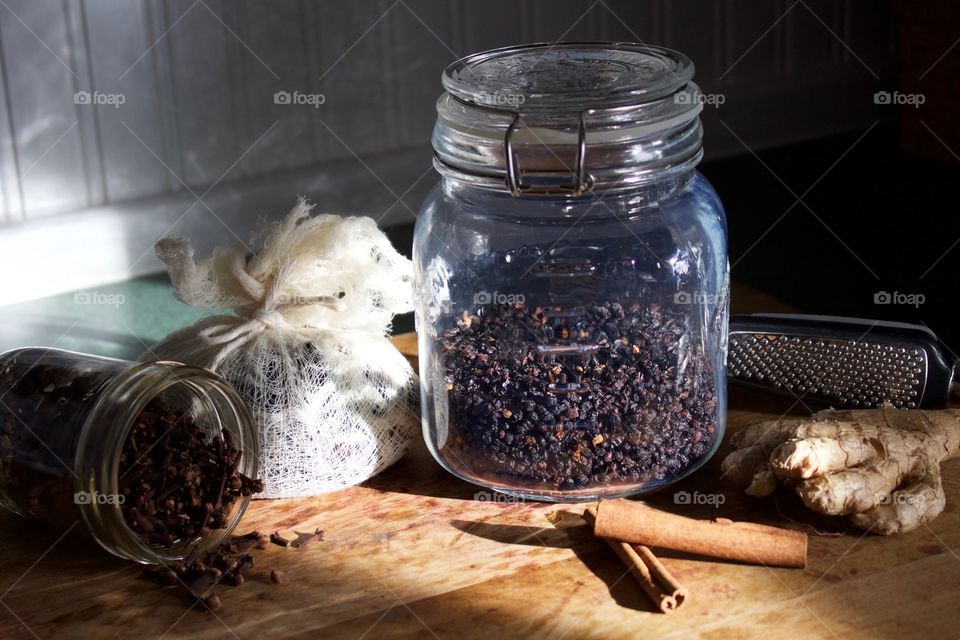 Ingredients for elderberry syrup—cloves in small glass jar, elderberries and chopped ginger in cheesecloth, dried elderberries in sealed glass jar, cinnamon sticks and ginger root with grater on wooden surface