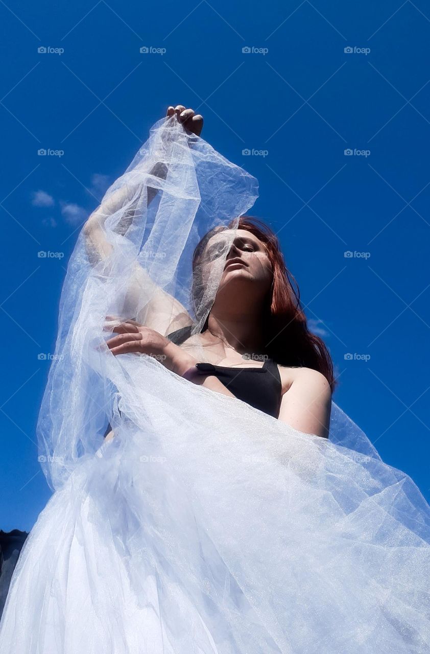 original portrait of a red-haired girl against the background of a clear, blue sky holding a light, white fabric, bottom view