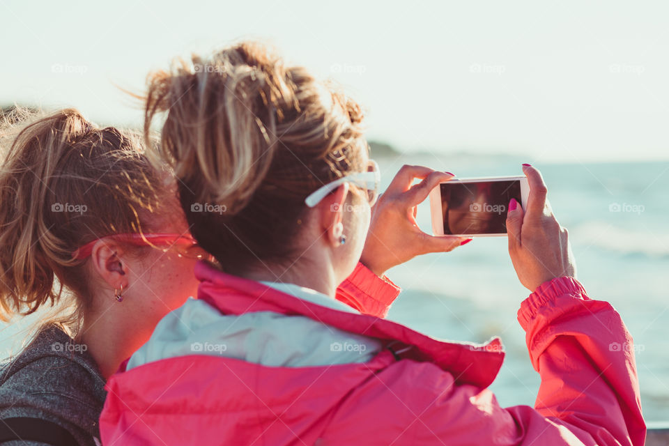 Young woman taking a photos using phone, looking at screen, standing outdoors, she is backlighted by sunlight with plain sky and sea in the background