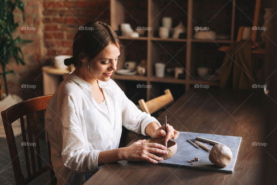 Young attractive woman in white shirt ceramic artist decorating clay plate with tool at table in pottery workshop. Handmade work student, freelance small business