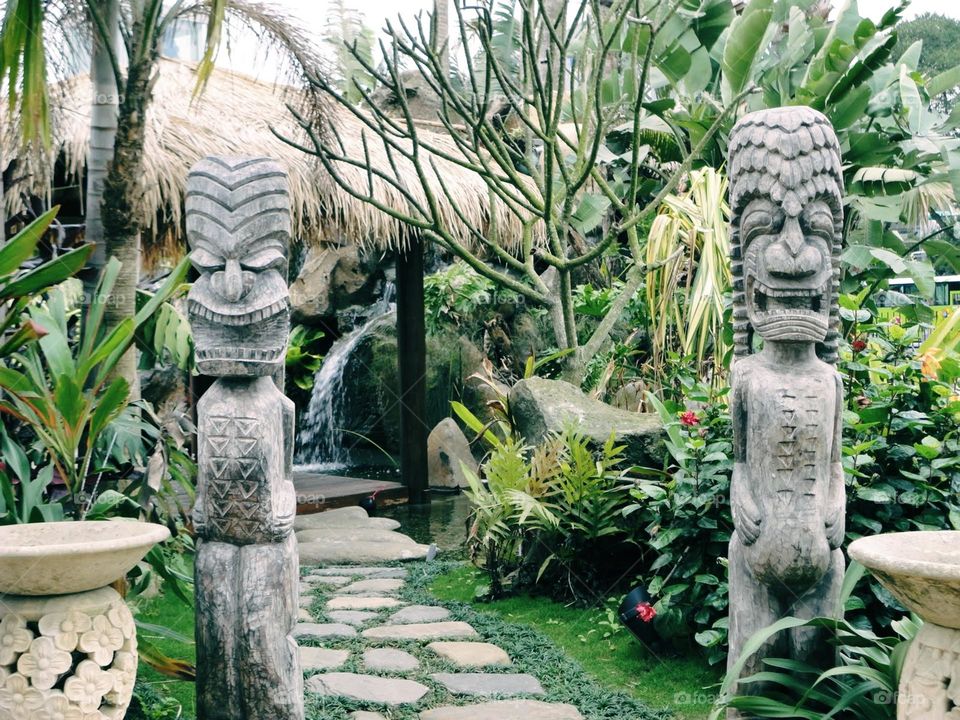 A dreamlike garden with a winding path leading to a quaint hut adorned with intricate stone carvings and a thatched roof. A gentle waterfall flows in the background, surrounded by lush tropical plants, creating a serene and enchanting atmosphere.