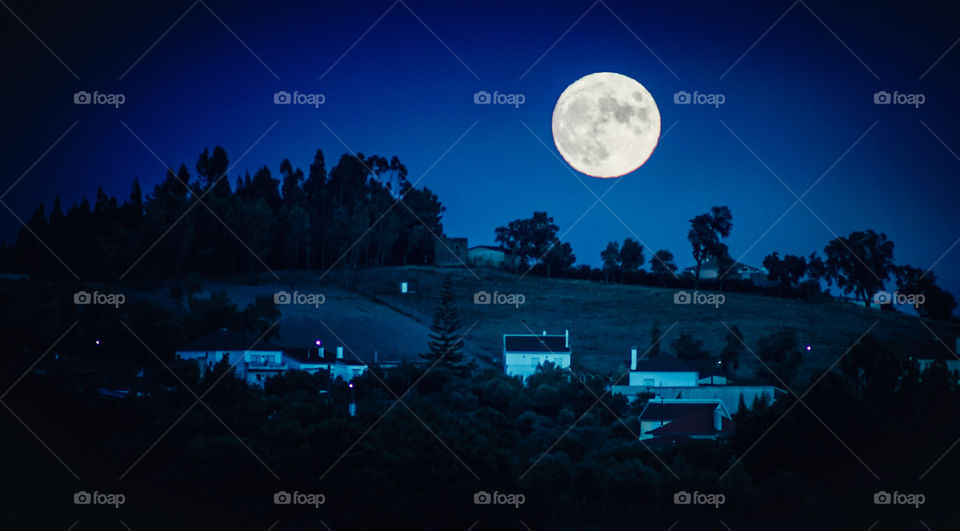 The moon is large, full and low in the sky as it rises behind the hill which is dotted with white cottage and lots of trees, in Central Portugal 