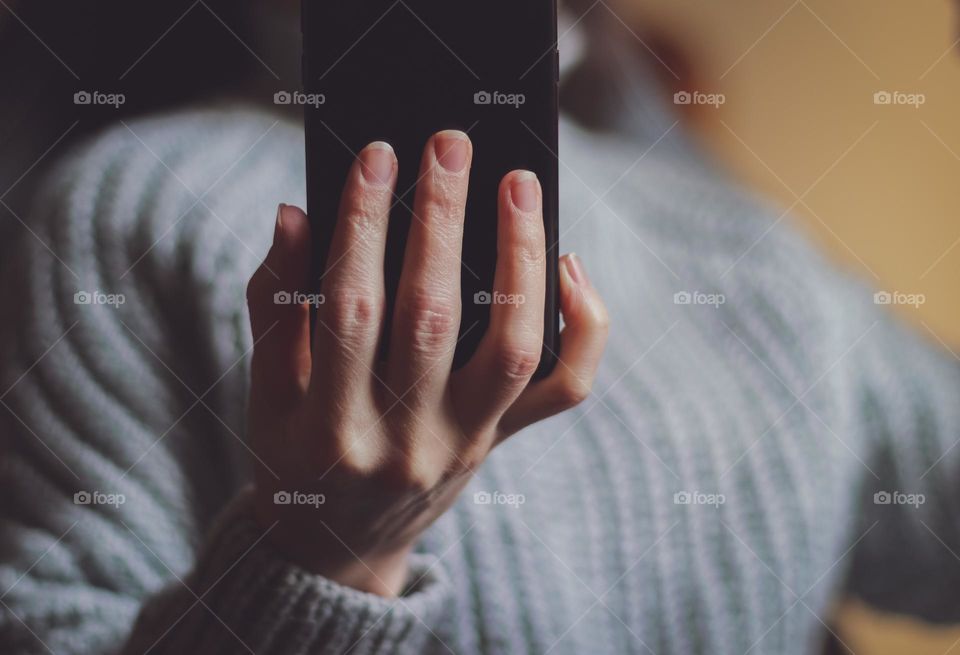 The hand of a young caucasian girl in a gray knitted sweater holds a black mobile phone in front of her while chatting with her friend on social networks, side view close-up in dark style.