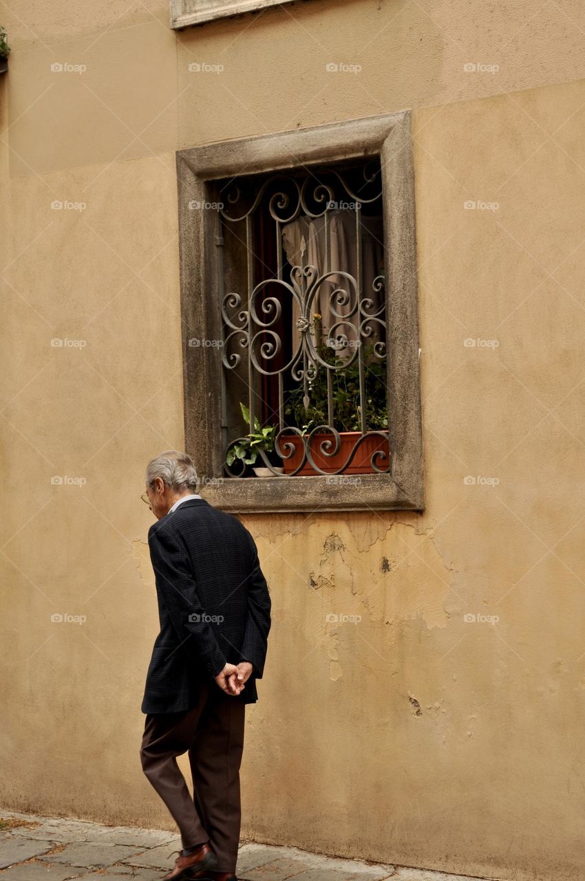 Elderly man walking on the street passing by a yellow painted wall and window decorated with plants and railings 