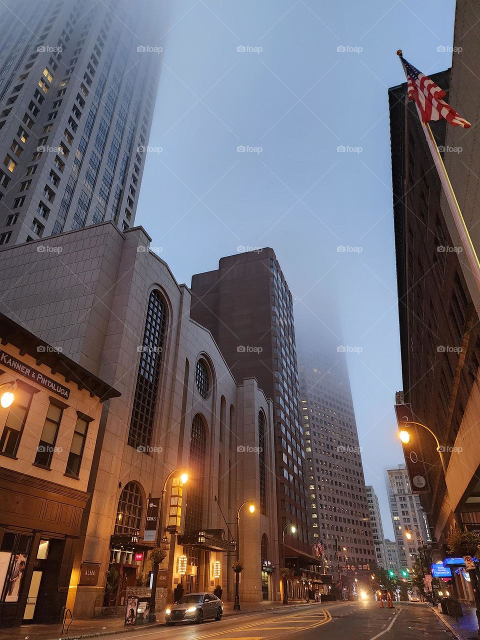 Tall buildings are shrouded in fog on a warm and humid summer morning while lights cast a warm glow