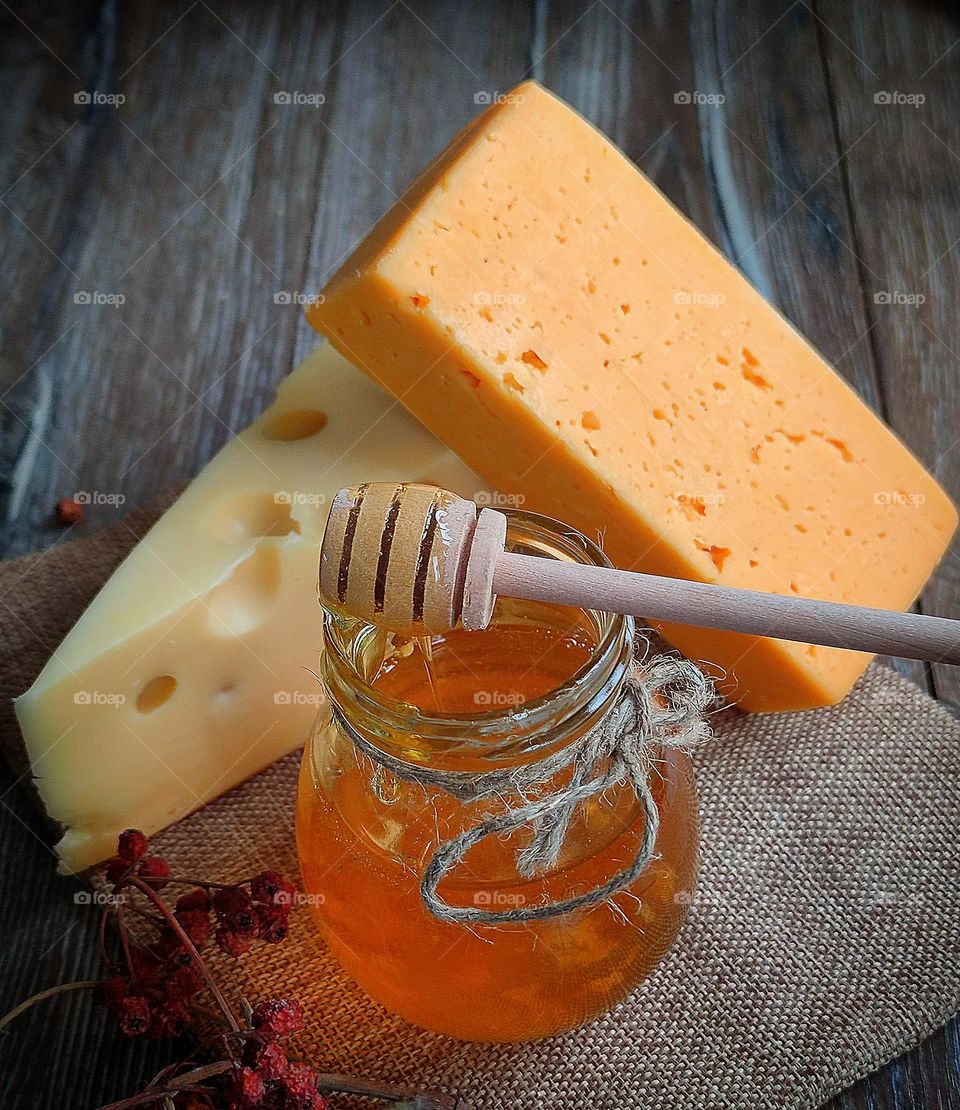 Food. On a wooden background lies burlap, on which lie two types of cheese: white "Masdam" and yellow "Cheddar". In the foreground is a jar of honey on which lies a spiral wooden spoon. Honey is dripping from the spoon.