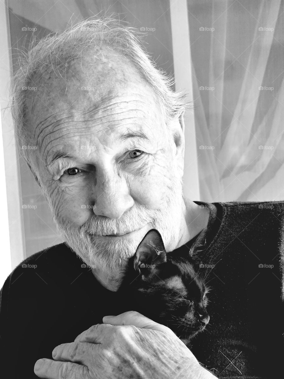 Black and white portrait of man holding a small black kitten. Man is smiling. He is looking straight ahead. The kittens eyes are almost closed. He is relaxed and happy to be held.