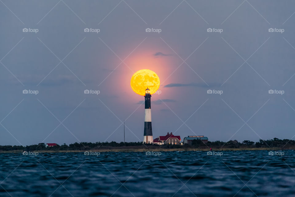 Full moon rising behind a tall lighthouse guarding the coastline, seen from across a body of water. 