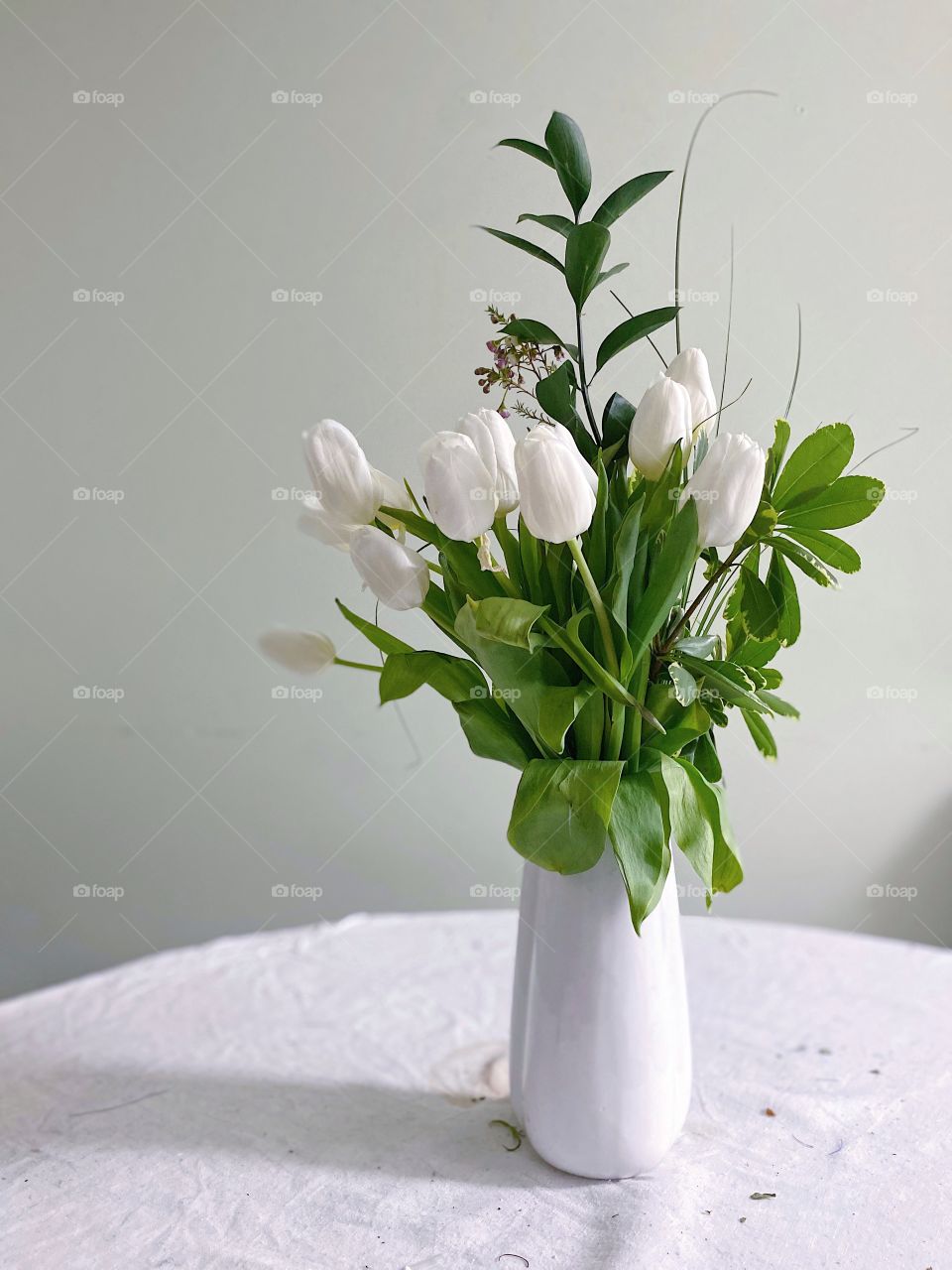 A white vase with white tulips and greenery atop a white linen table cloth.