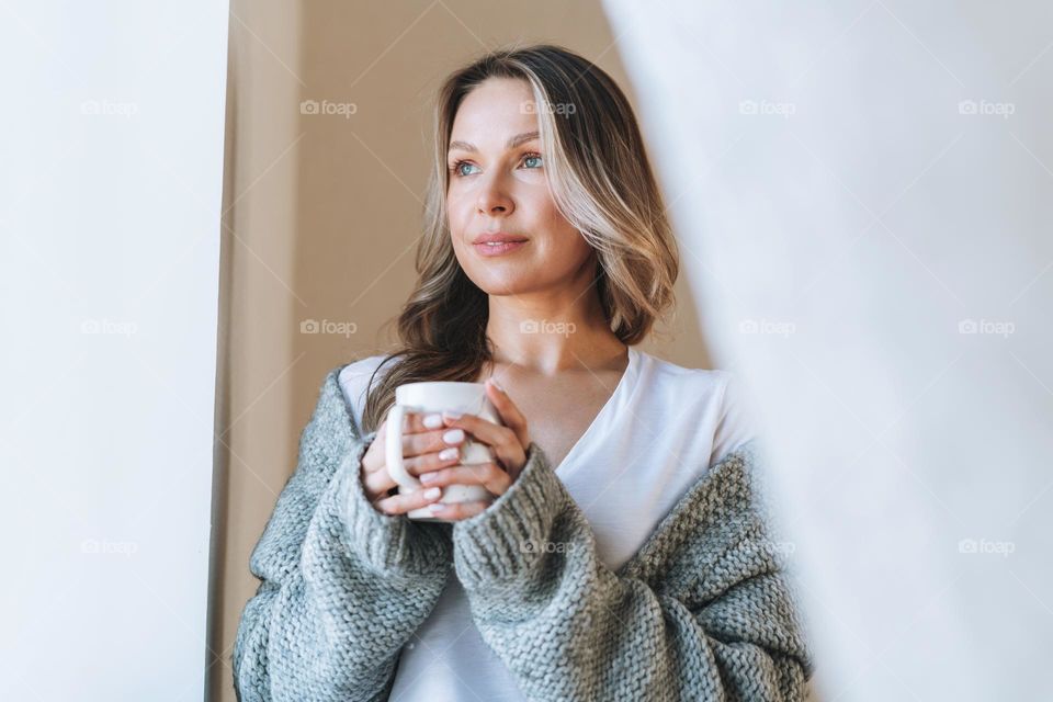 Young beautiful woman forty year with blonde long curly hair in cozy knitted grey sweater with cup of tea in hands in bright interior at the home