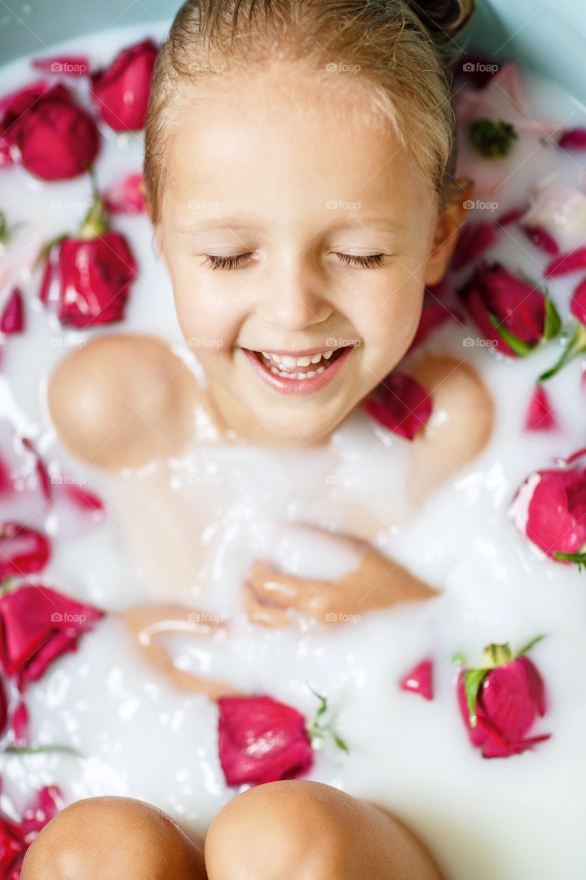 Cute little girl with blonde hair in milk bathroom with fresh red roses 