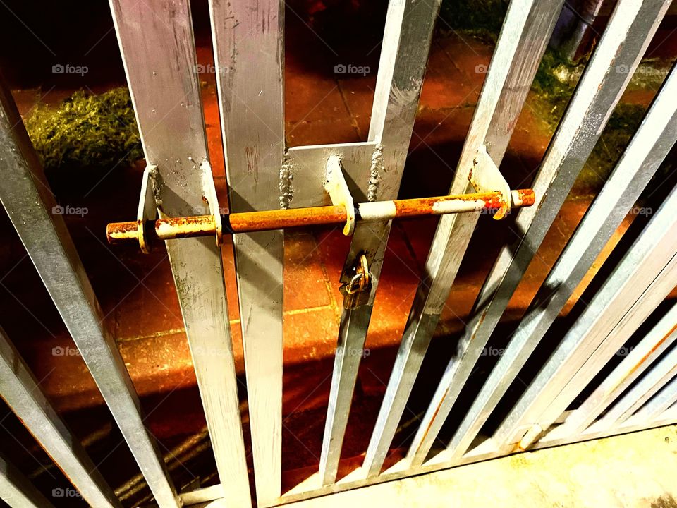 The lock to a gate. The white metal fence is locked by a simple padlock. Behind the dense is orange stone. 