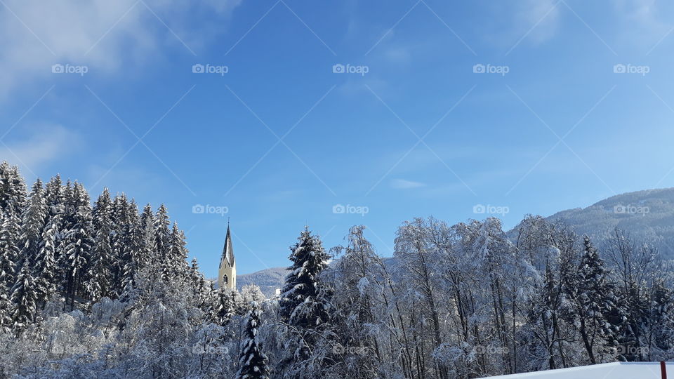 Winter in Austria. Church belltower surrounded by the woods. Blue Sky and snow.