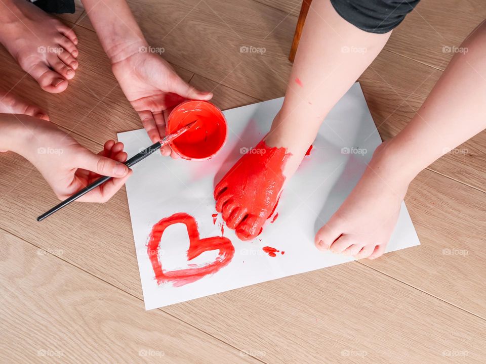 Two caucasian girls sisters paint their feet with red paint with a brush sitting on a wooden floor, side view close-up. Concept kids creative creativity, hobby, body painting, diy.