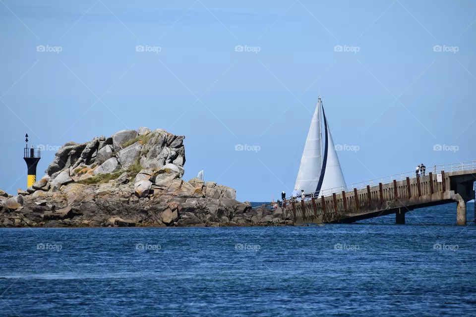 A ship with a sail near  sea and a bridge 