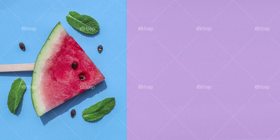 One sliced ​​rectangular shaped watermelon on a wooden stick with mint leaves and pits lies on the left on a lilac blue background with copy space on the right, flat lay close-up. End summer treat, summer food, watermelon ice cream, natural food, hea