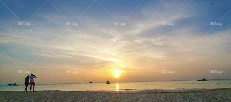 sunset over the ocean on a tropical island with boats in the water and a loving couple admiring the view from the sand.