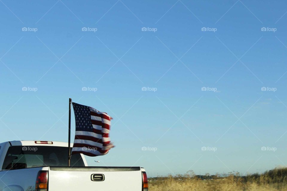 an American flag flapping in the Wind on the back of a white pickup truck driving on the freeway