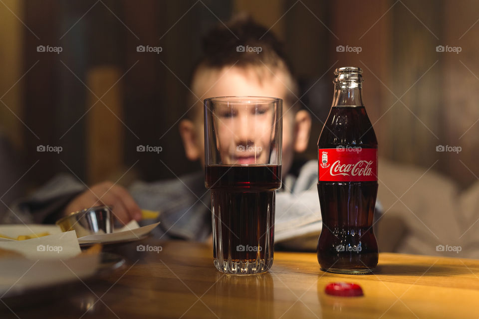 a cheerful, cute guy of fifteen years old, sitting in a cafe, smiling, having fun and looking through two bottles of Coca-Cola. dressed in denim, charismatic teen