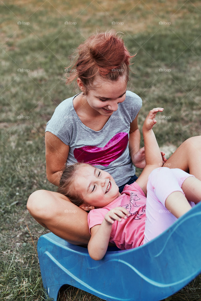 Teenage girl playing with her younger sister in a home playground in a backyard. Happy smiling sisters having fun on a slide together on summer day. Real people, authentic situations