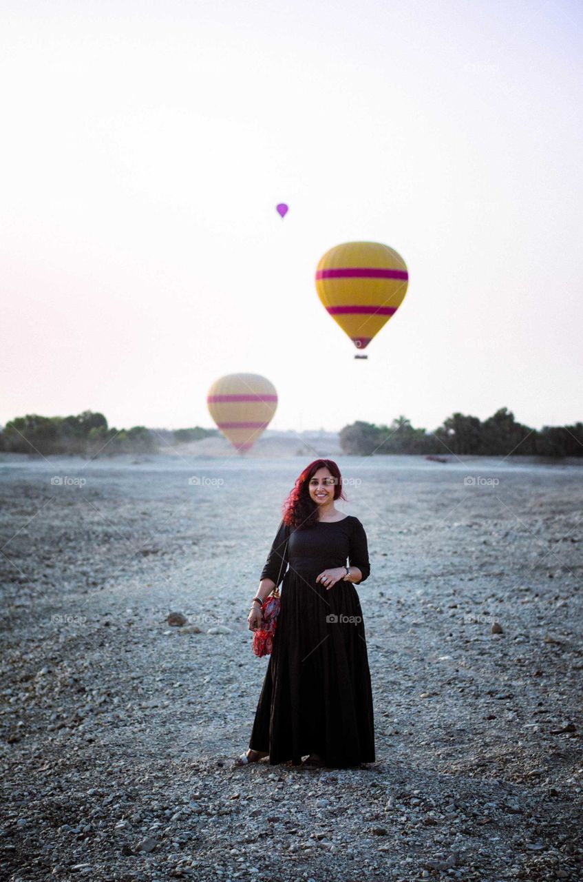 Girl smiling in the desert in long dress with Hot Air Balloons in the air behind her
