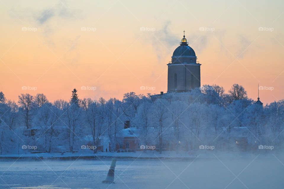 Arrival by ferry to the Suomenlinna fortress island on an extremely cold winter morning at sunrise with sea fog and icy Baltic sea, the Suomenlinna church (Suomenlinnan kirkko) & lighthouse and historical barracks behind frosty trees in the backgroun