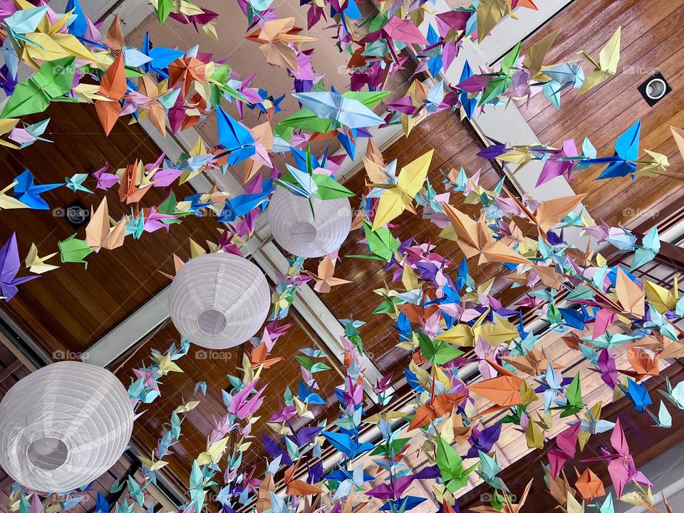 An impressive decoration of colorful origami birds hanging in a modern environment, with white lanterns in the background. The union of vibrant colors and the smooth movement of the papers create a cheerful and playful atmosphere.