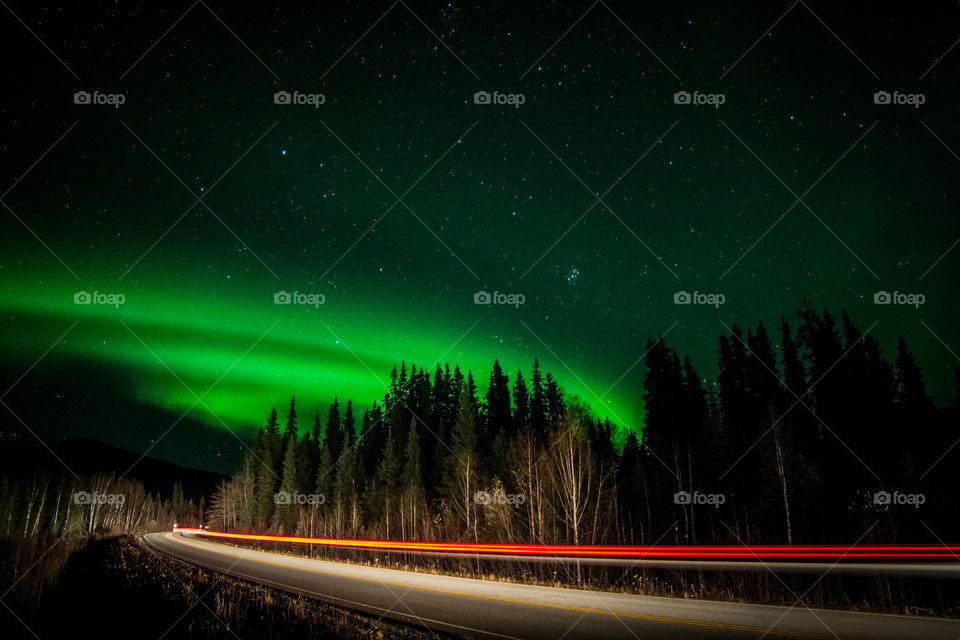 The aurora borealis streaks across the sky as a car does the same on the road below.
