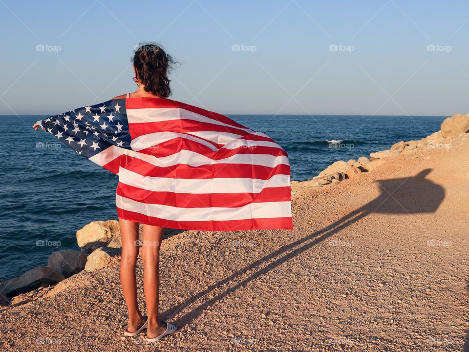 A Caucasian teenage girl with an American flag on her back is standing on the beach at sunset, close-up view from the side.