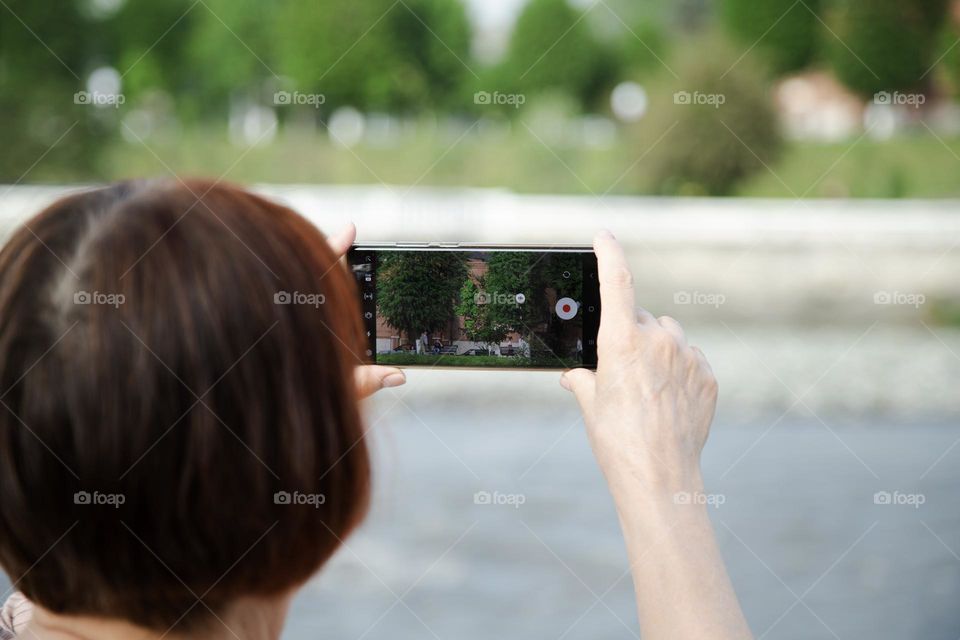 Caucasian woman taking a video with a cell phone camera, standing on the river bank at sunny summer day . Video creator concept .