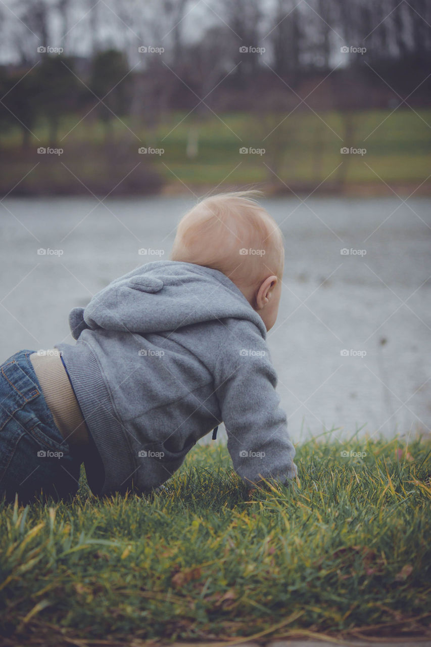 Baby crawling in jeans and hooded sweatshirt outside in grass near water or lake