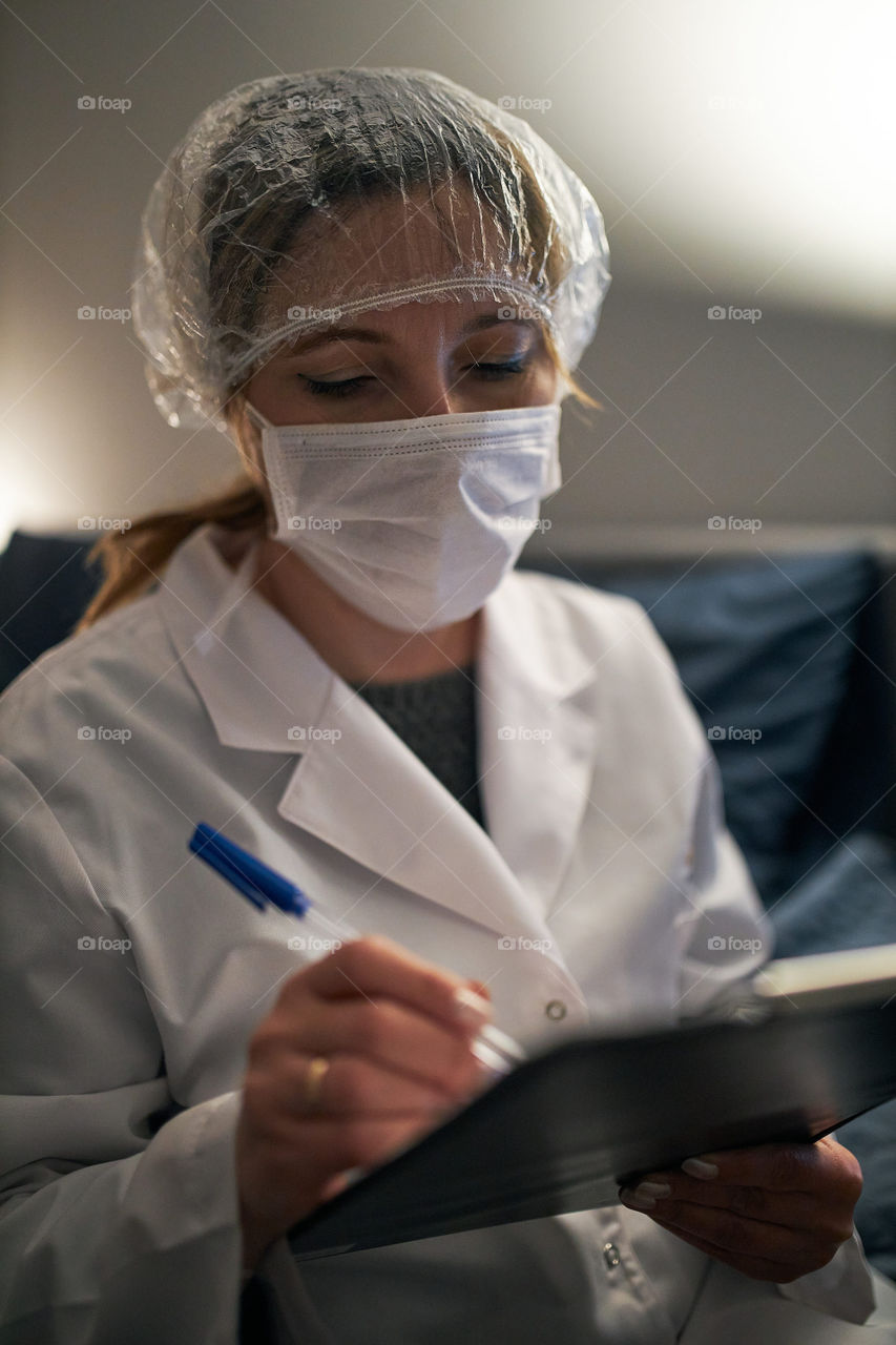 Doctor filling out a document. Hospital staff working at night duty. Woman wearing uniform, cap and face mask to prevent virus infection