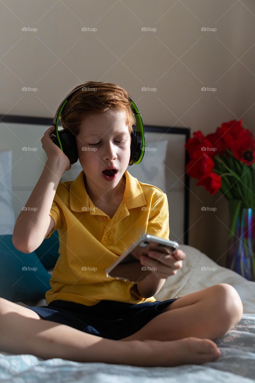 Red-haired child boy listens to music in headphones and holds a phone in his hands