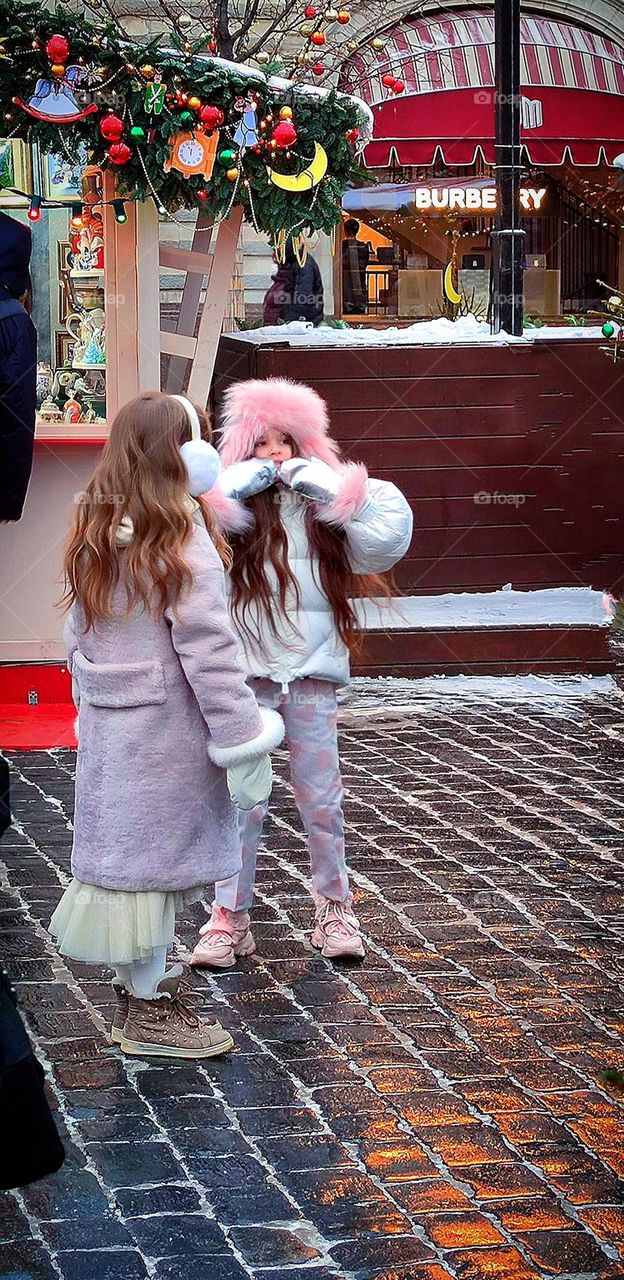 Happiness.  On the winter street there are two girls who are warmly dressed.  One girl smiles and shows something and tells her friend