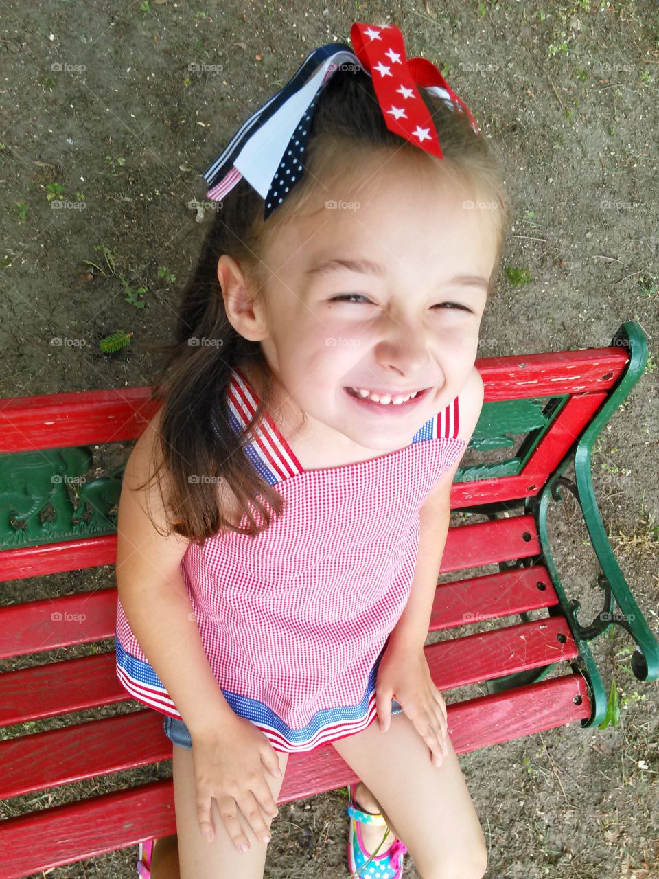 American Girk. Little girl dressed in patriotic clothing smiling on a small red bench. 