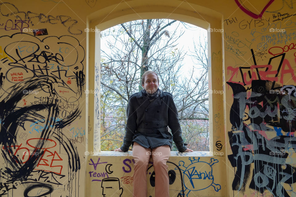 A young man in a coat sitting at the window in a building with graffiti.