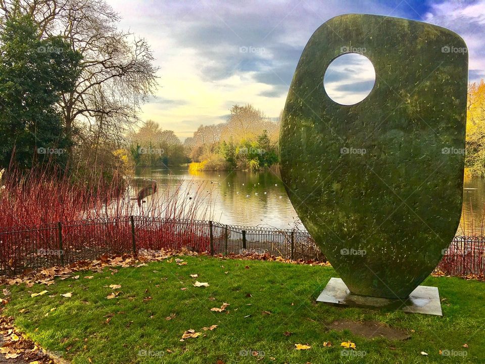 A gorgeous abstract sculpture in a park in London. The background shows a fall scene: leave-less trees, vibrant red shrubs and a reflective pond. 