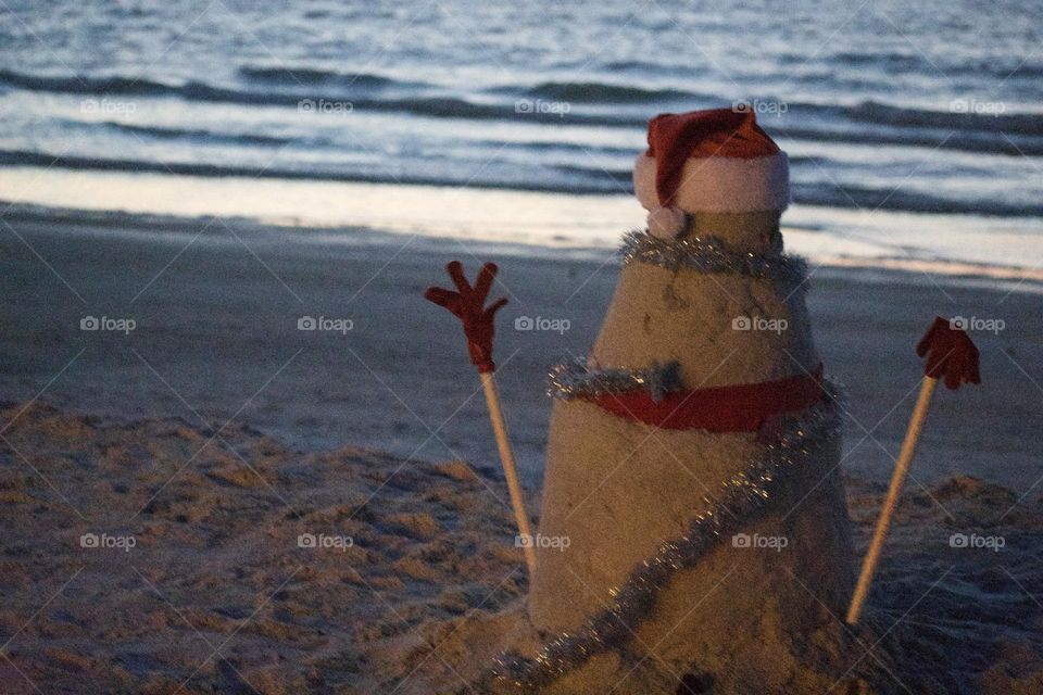 A sand figure on the beach wears a Santa hat and has two sticks on either side of its body to represent arms. The figure has the appearance of a snowman, except made out of sand.