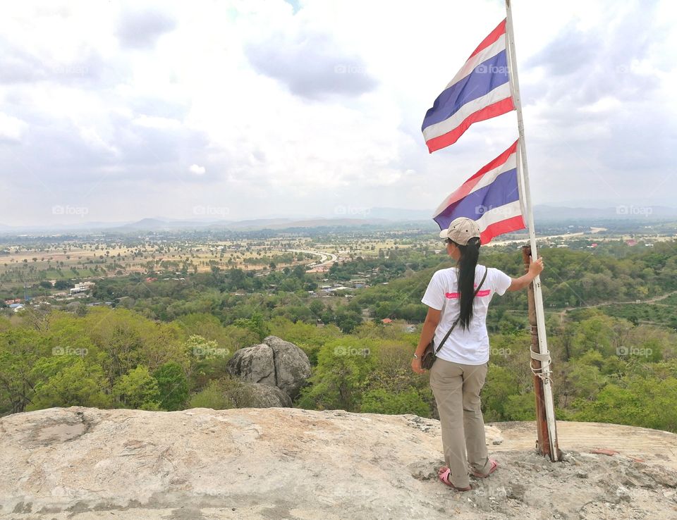 An Asian girl hold the Thailand flag pole at the mountain peak.