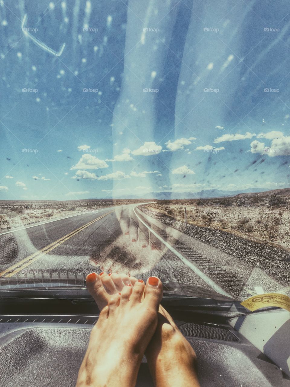 Female feet with red toenails resting on the dashboard of the passenger side of a car, driving along the open road in Death Valley desert. The road is long and empty 