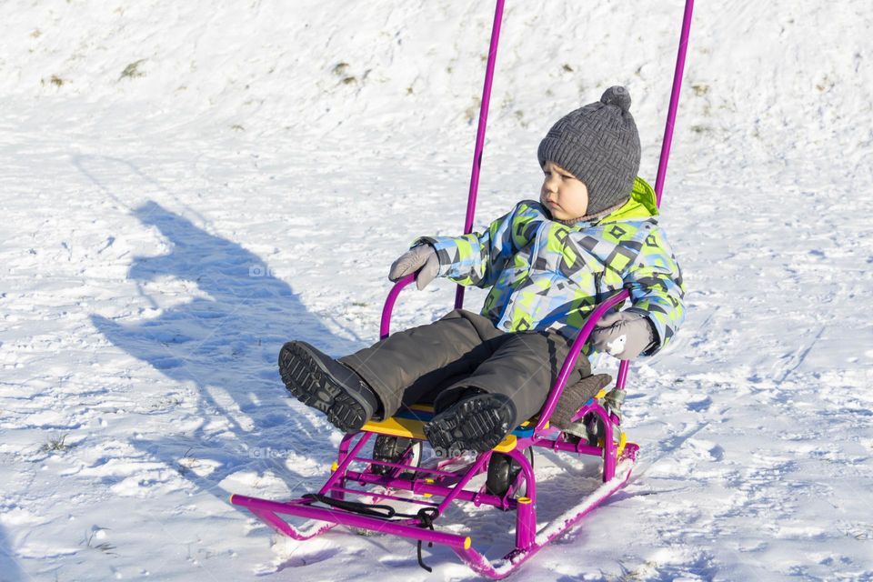 A child in winter in winter jackets, pants, a hat and boots on white snow on the street and in the park in nature plays winter fun and sleds.