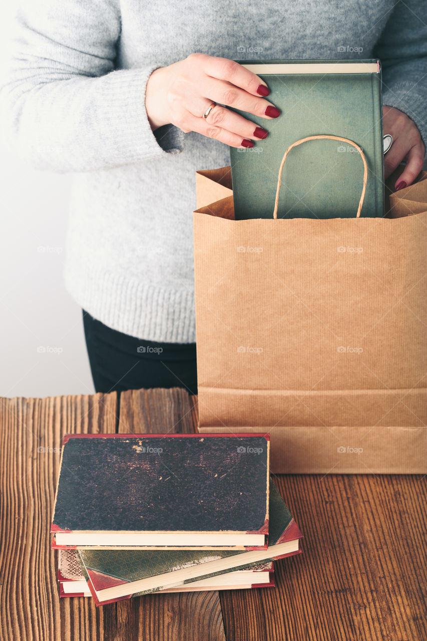 Young woman putting old books to paper bag in antique bookstore. Woman wearing grey sweater and jeans. Vertical photo