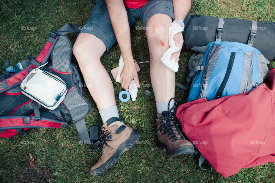 Woman injured in the accident on mountain hike. Dressing the wound on her knee with medicine in spray and gauze sitting on grass. Equipped with backpack wearing sportswear