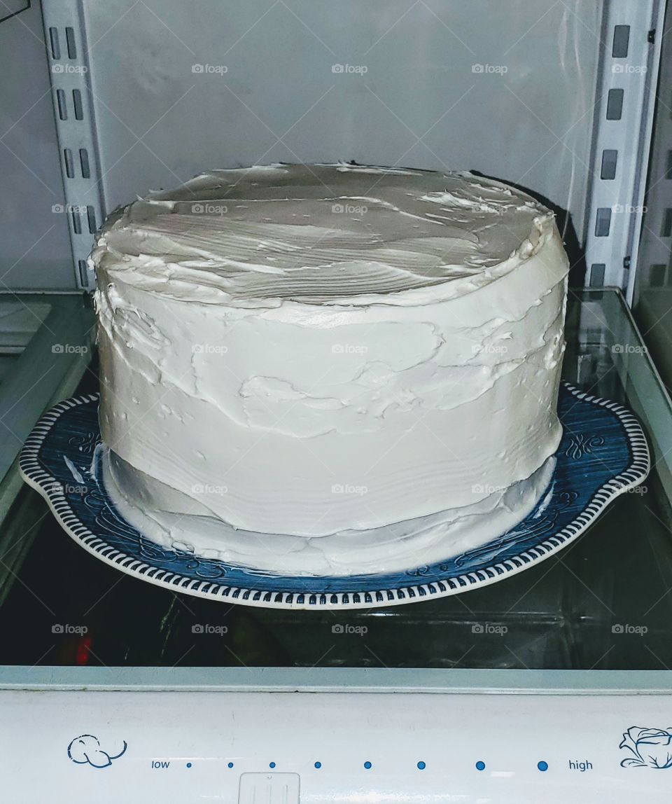 Cake with white frosting on refrigerator shelf, ready for more decorating. This is going to be a birthday cake.