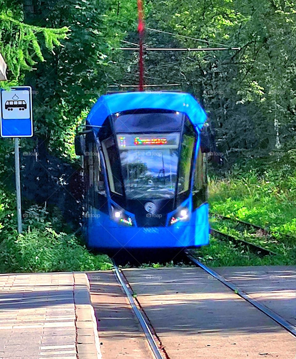 Public transport.  A blue tram exits a patch of trees onto a landing platform.