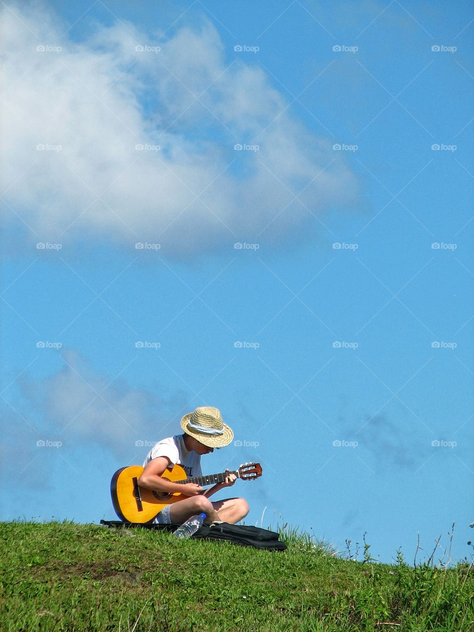 A young boy plays his guitar on a hillside with a beautiful blue sky overhead.