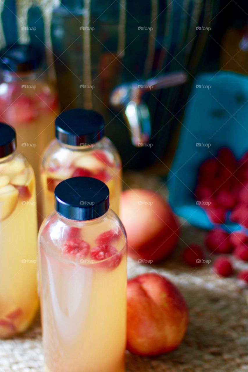 Kombucha, bottled for a second-ferment, flavored with nectarines, raspberries and ginger root slices on a natural fiber mat, whole nectarines and raspberries in a paper carton, stoneware kombucha crock with a stainless steel spigot in the background